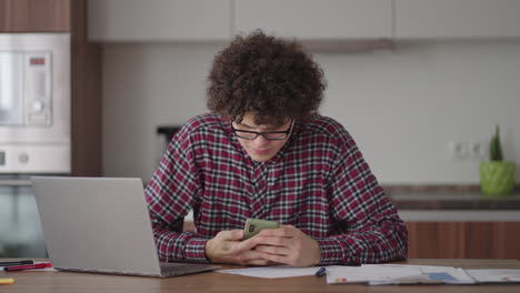 Curly-haired-Male-student-attractive-young-boy-in-glasses-is-studying-at-home-using-laptop-typing-writing-in-notebook.-College-student-using-laptop-computer-watching-distance-online-learning-seminar